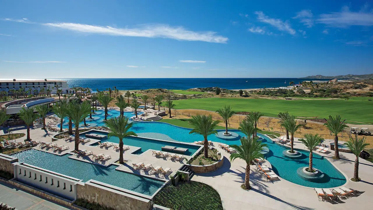 Aerial view of a luxury resort pool complex with palm trees, golf course, and ocean beyond