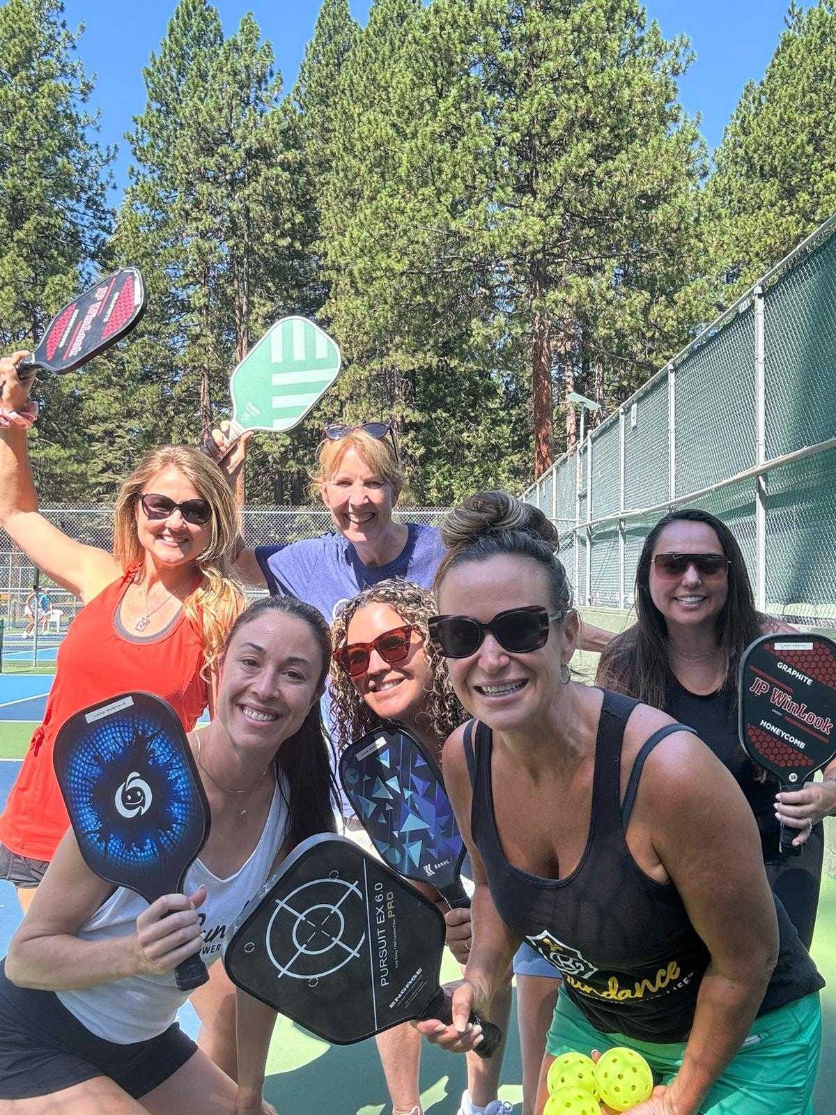 Six women smiling and holding pickleball paddles on an outdoor court surrounded by pine trees