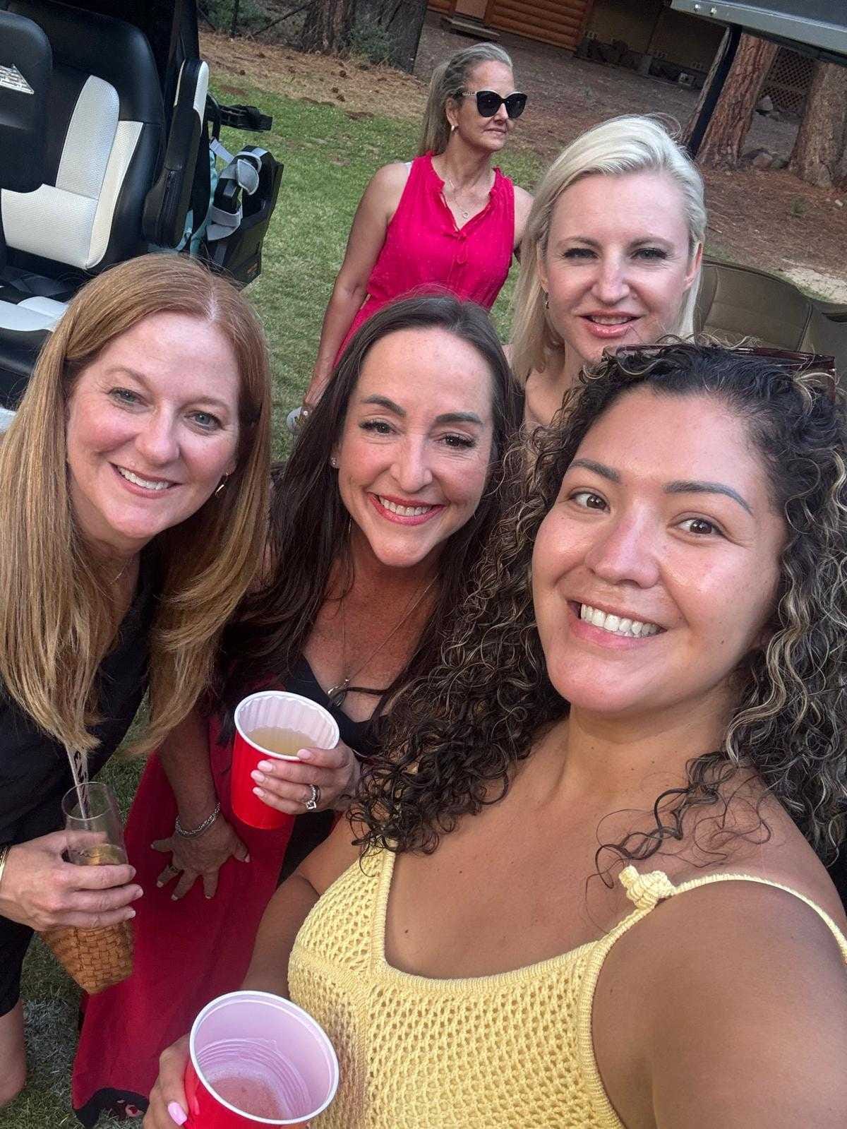 Five women smiling together at an outdoor gathering, holding drinks near trees